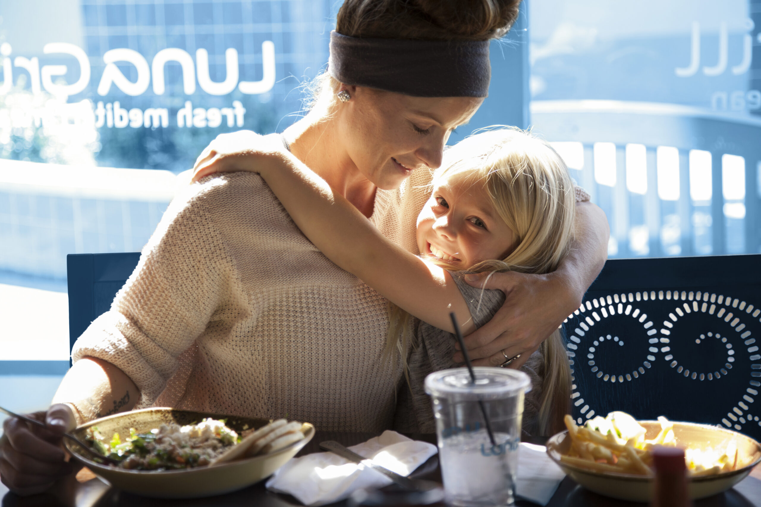A woman and a young girl are sitting at a table in a restaurant, sharing a warm hug and smiling at each other, with plates of food in front of them.